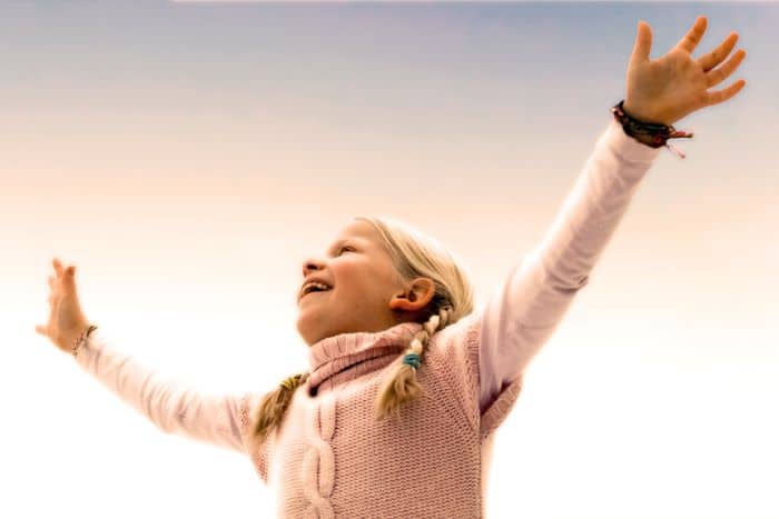 A smiling child in pink sweater with open arms facing the sky wearing a colorful mindfulness bracelet.