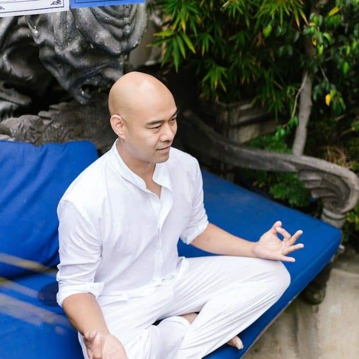 A man dressed in white on a meditation mat and cushion.